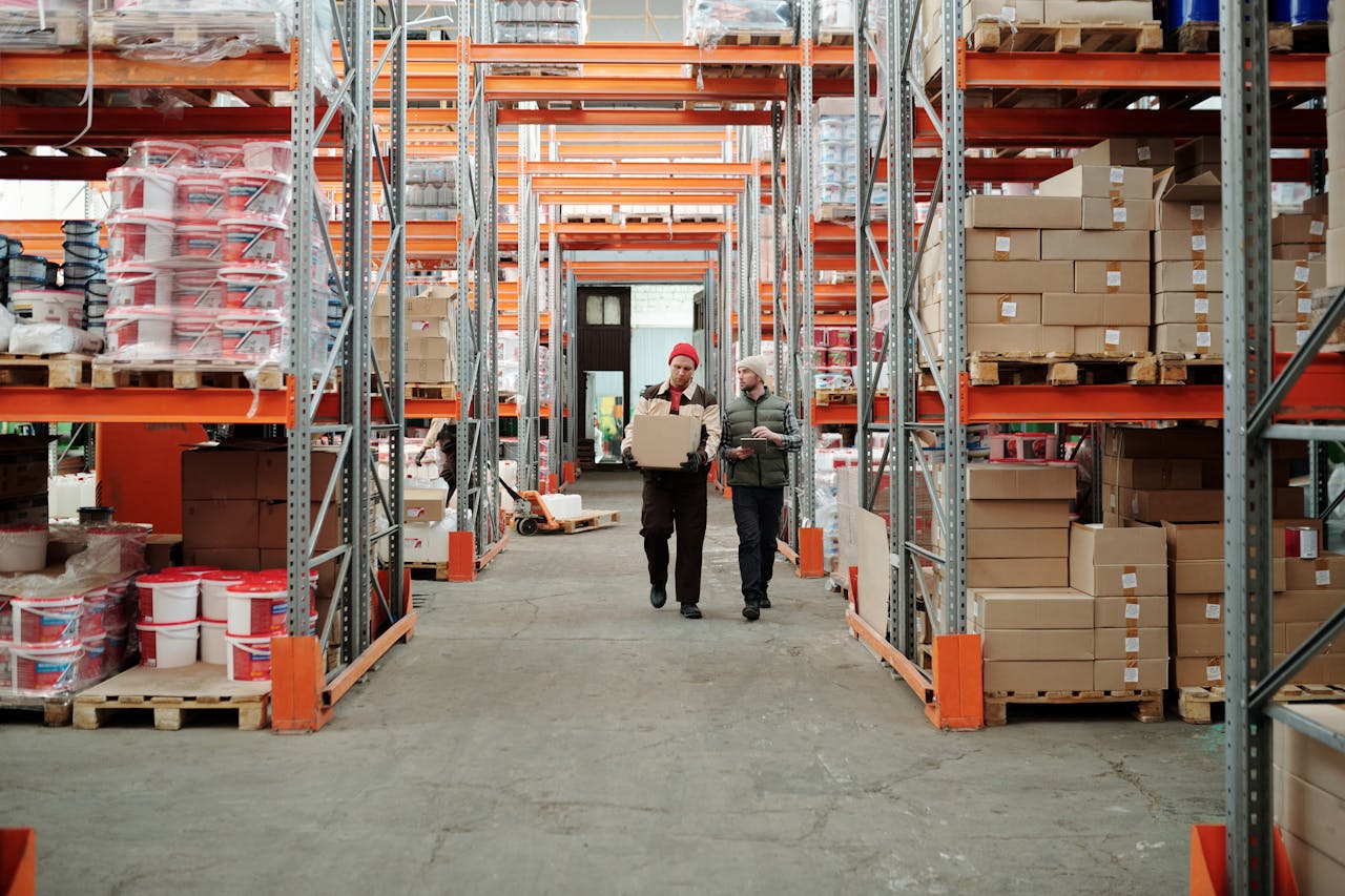 gallery-04 Two workers handle a package in a spacious warehouse surrounded by shelves stocked with boxes and products.